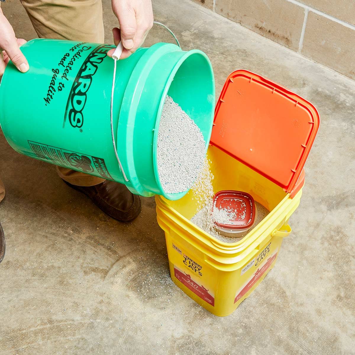 A green bucket is being tilted to pour its contents into a yellow bucket, which is sitting on a concrete floor, with an orange lid partially closed.