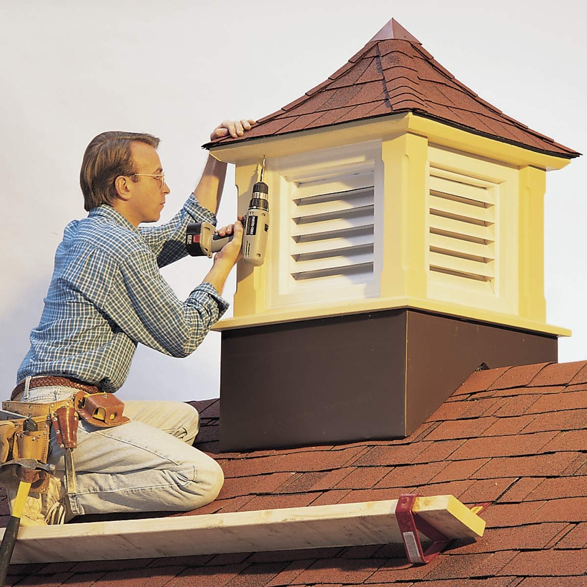 A man is using a power drill to install a vent on a rooftop, surrounded by brown shingles and a supportive wooden plank.