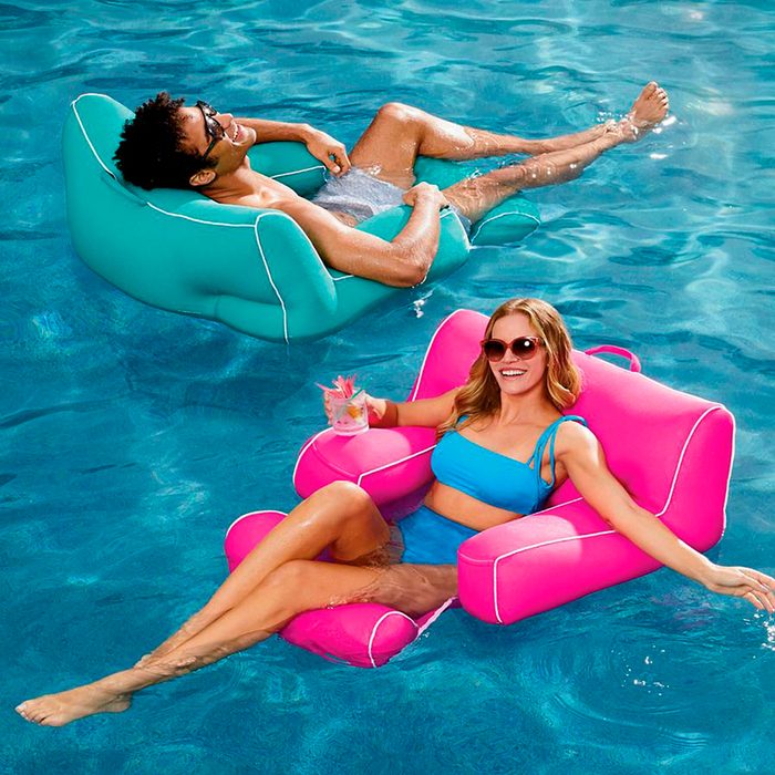 Two individuals relax on colorful floating chairs in a pool, one sipping a drink, enjoying the sunny environment.
