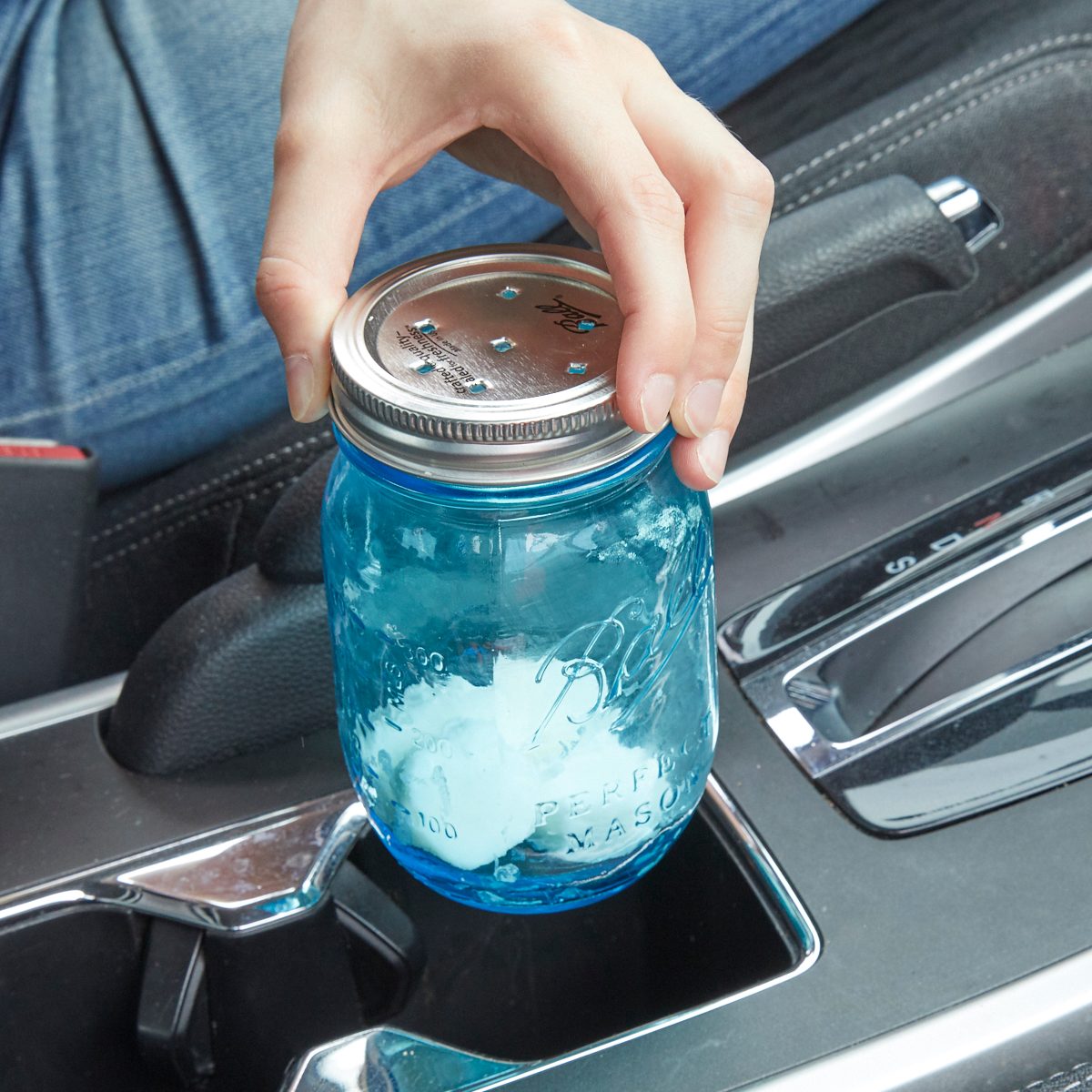 A hand lifts a blue mason jar from a car's center console, which contains ice. The vehicle's interior suggests a casual setting.