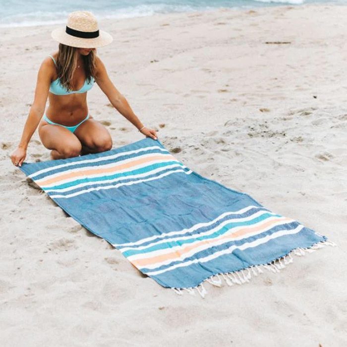 A woman in a bikini kneels on sandy beach, spreading out a striped towel while near the ocean under a sunny sky.