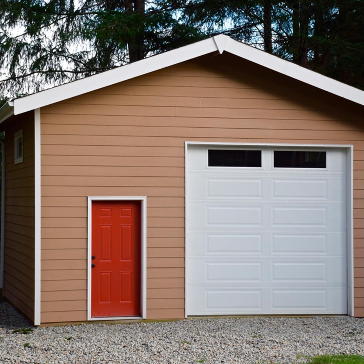 A brown shed features a bright red door and a white garage door. Pebbles cover the ground, with trees visible in the background.