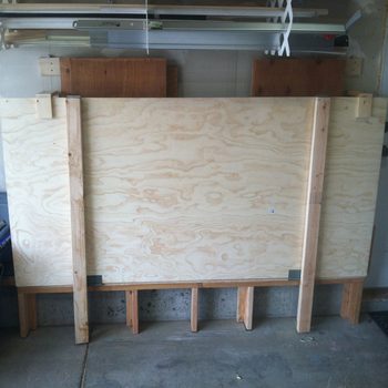 A large, flat plywood board is supported by wooden beams, positioned in a garage environment with scattered tools and materials in the background.