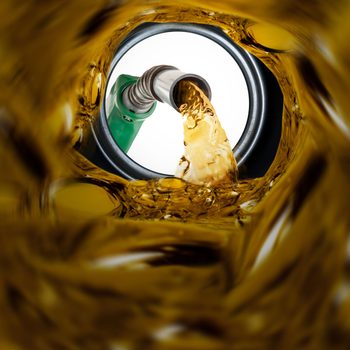 A fuel nozzle pours liquid gasoline into a tank, surrounded by a swirling golden liquid in a bright environment.