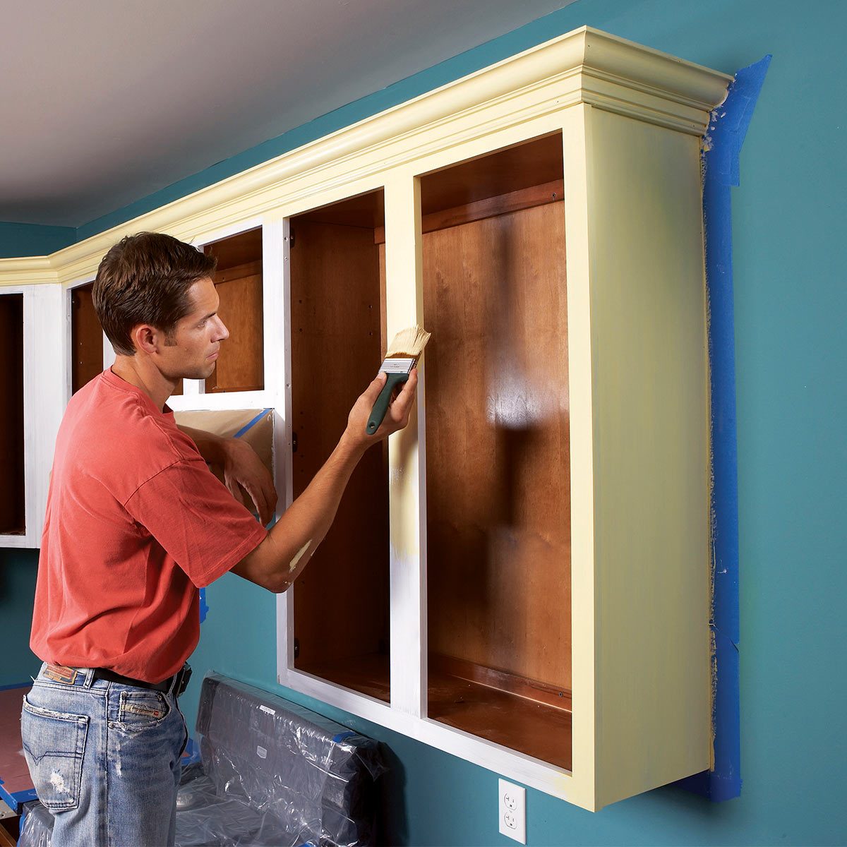 A man paints a yellow cabinet in a room with teal walls, while protecting edges with blue tape. Plastic covers nearby furniture.