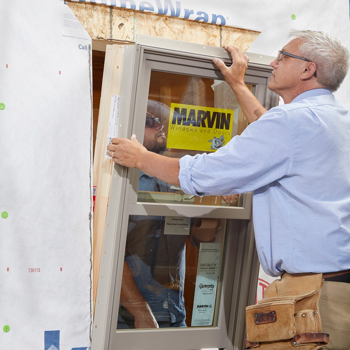 A worker installs a window frame in a partially constructed building, with protective wrapping and materials visible in the background.