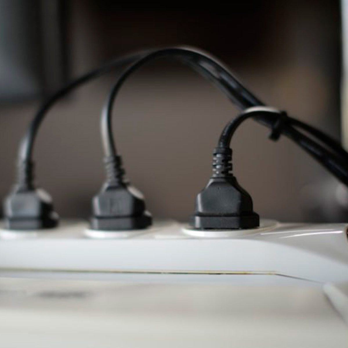 Three black power cords are plugged into a white power strip, set against a blurred, neutral-colored background.