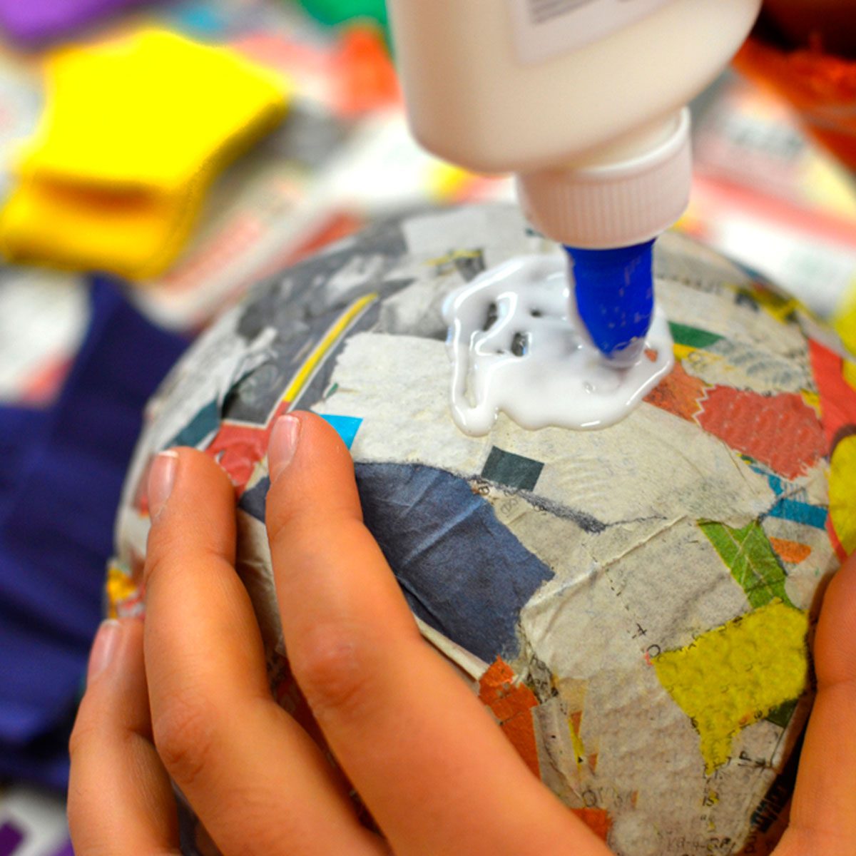 A hand applies glue to a paper-mâché ball, surrounded by colorful craft materials on a table, indicating an art or crafting environment.