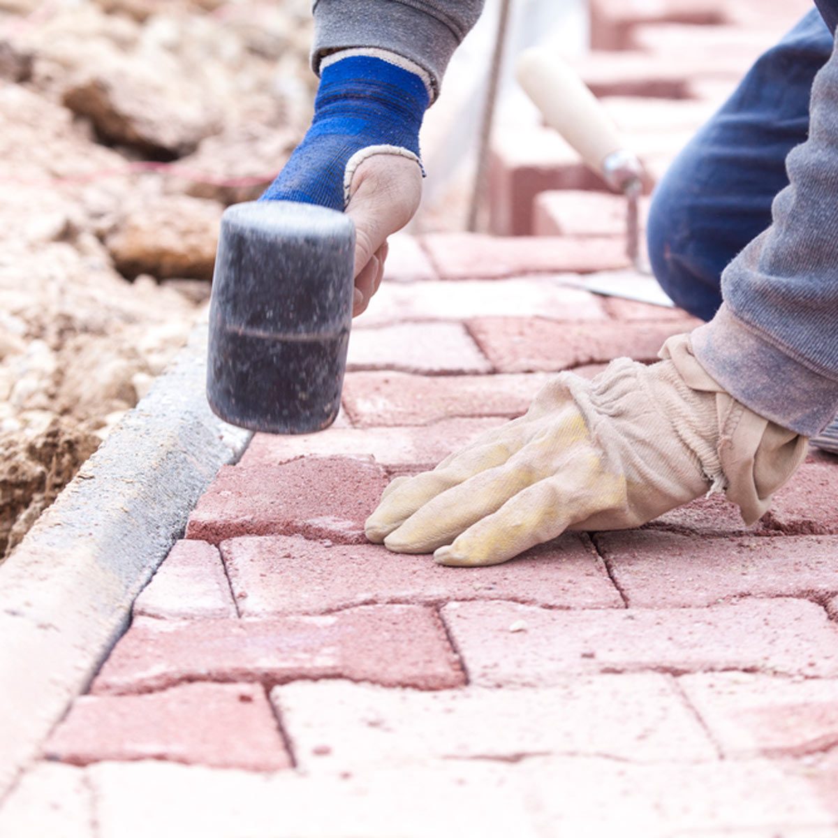A person's gloved hand uses a mallet to tap down brick pavers being laid on a pathway, surrounded by loose gravel and tools.
