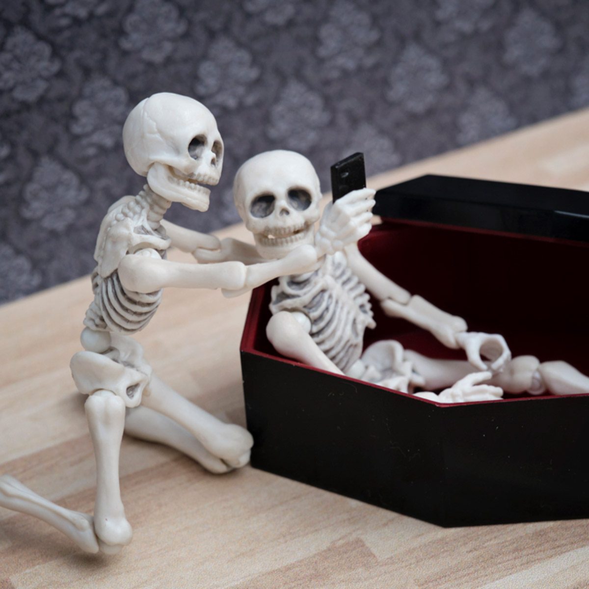 Two skeleton figures pose for a selfie while one leans out of a black coffin, set against a patterned backdrop on a wooden surface.