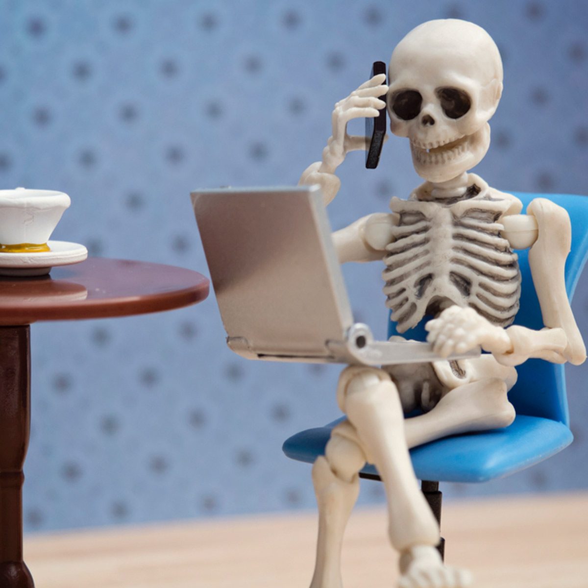 A skeleton sits at a blue chair, using a laptop while holding a phone to its ear, next to a table with a cup.