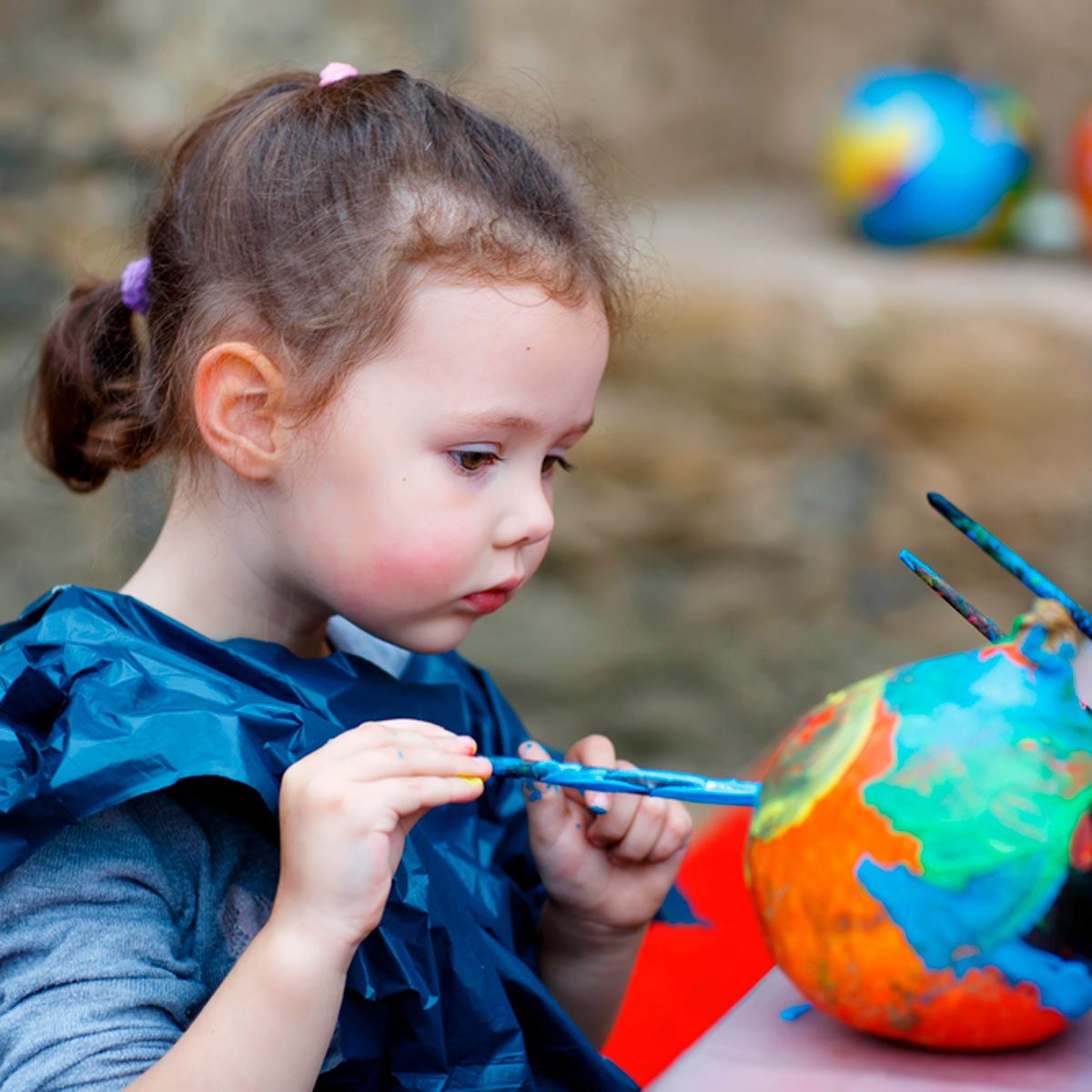 A girl paints a colorful globe with brushes while focused, wearing a blue smock; a blurred backdrop features more globes, suggesting a creative environment.