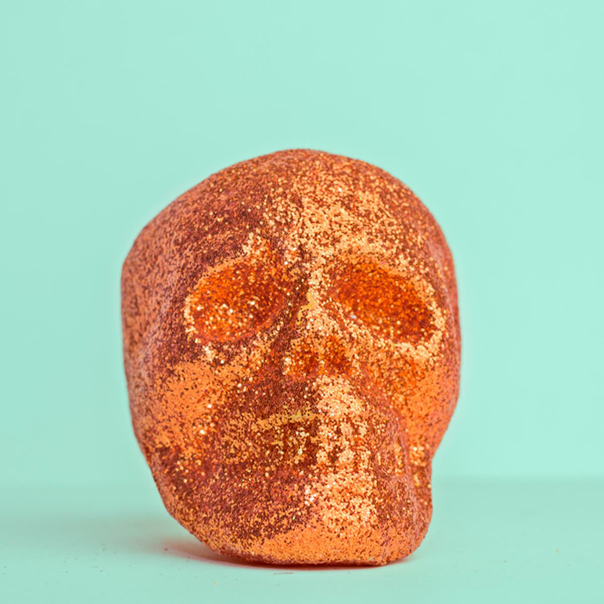 A glittery orange skull sits on a flat surface, contrasting against a soft turquoise background. Its surface sparkles under light, creating a whimsical effect.