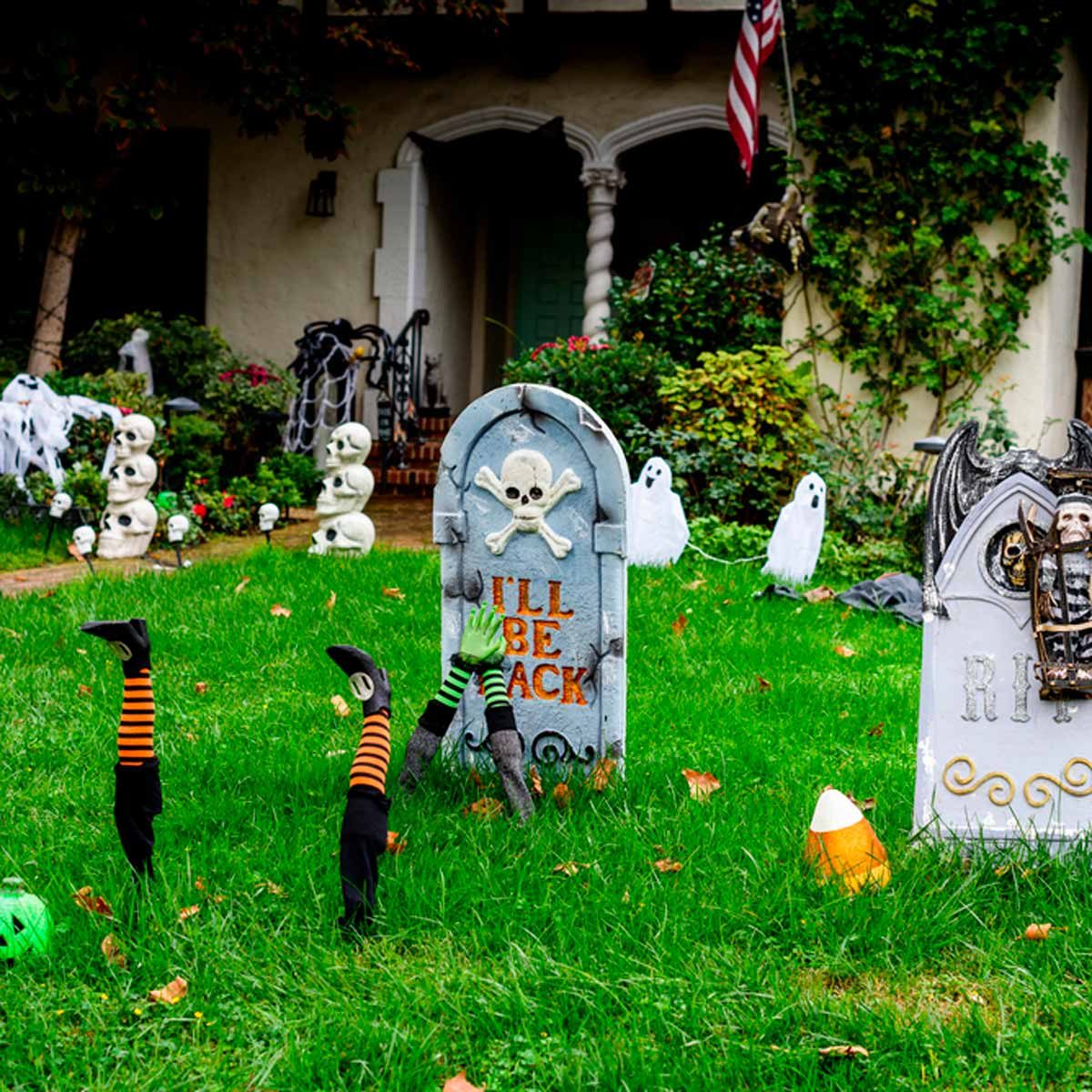 A gravestone with a skull displays "I'LL BE BACK." Nearby, striped arms emerge from the grass, surrounded by Halloween decorations like ghosts and skeletons.