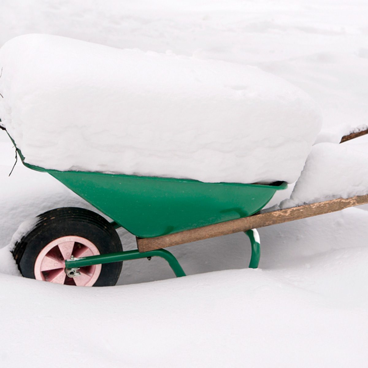 A green wheelbarrow sits partially buried under thick white snow, with only a portion of its frame and wheel visible amid the snowy landscape.