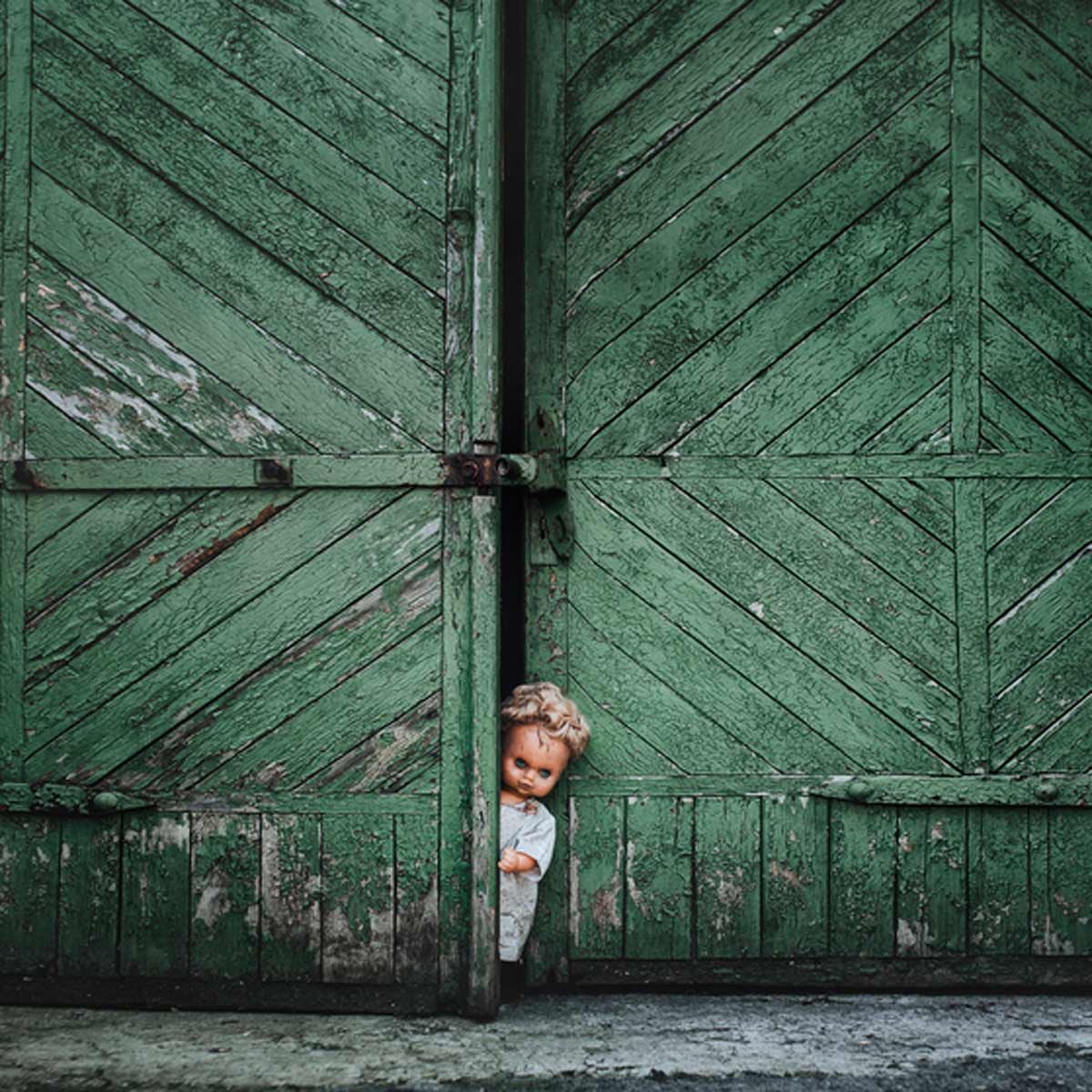 A vintage doll peeks out from a partially open pair of weathered green doors, set against a textured, aged wooden backdrop.