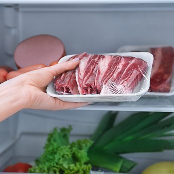A hand lifts a packaged tray of raw meat from an organized refrigerator, surrounded by lettuce, green onions, and various other food items.