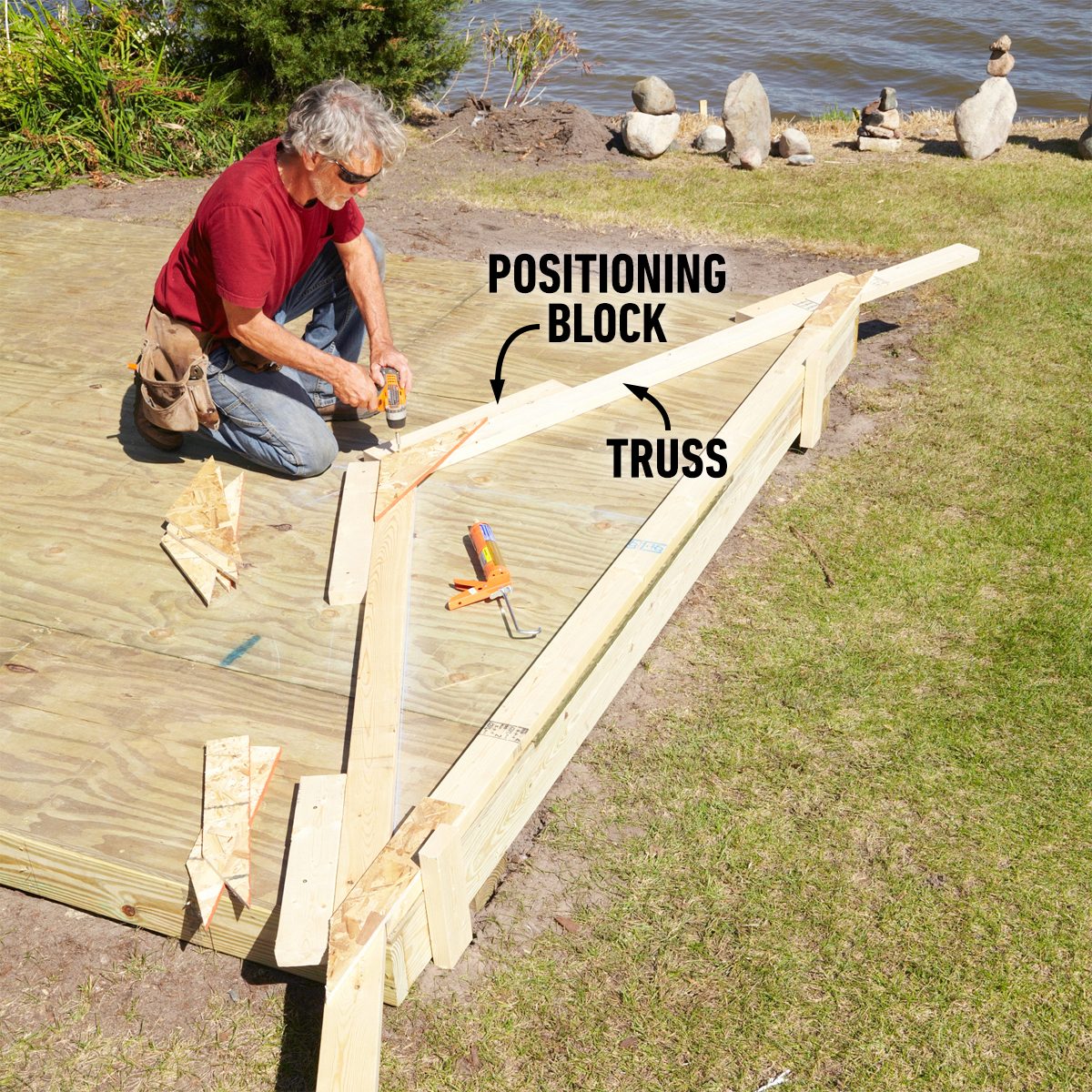 A man is securing a wooden truss onto a flat deck by using a drill, surrounded by tools and a tranquil lakeside environment.
