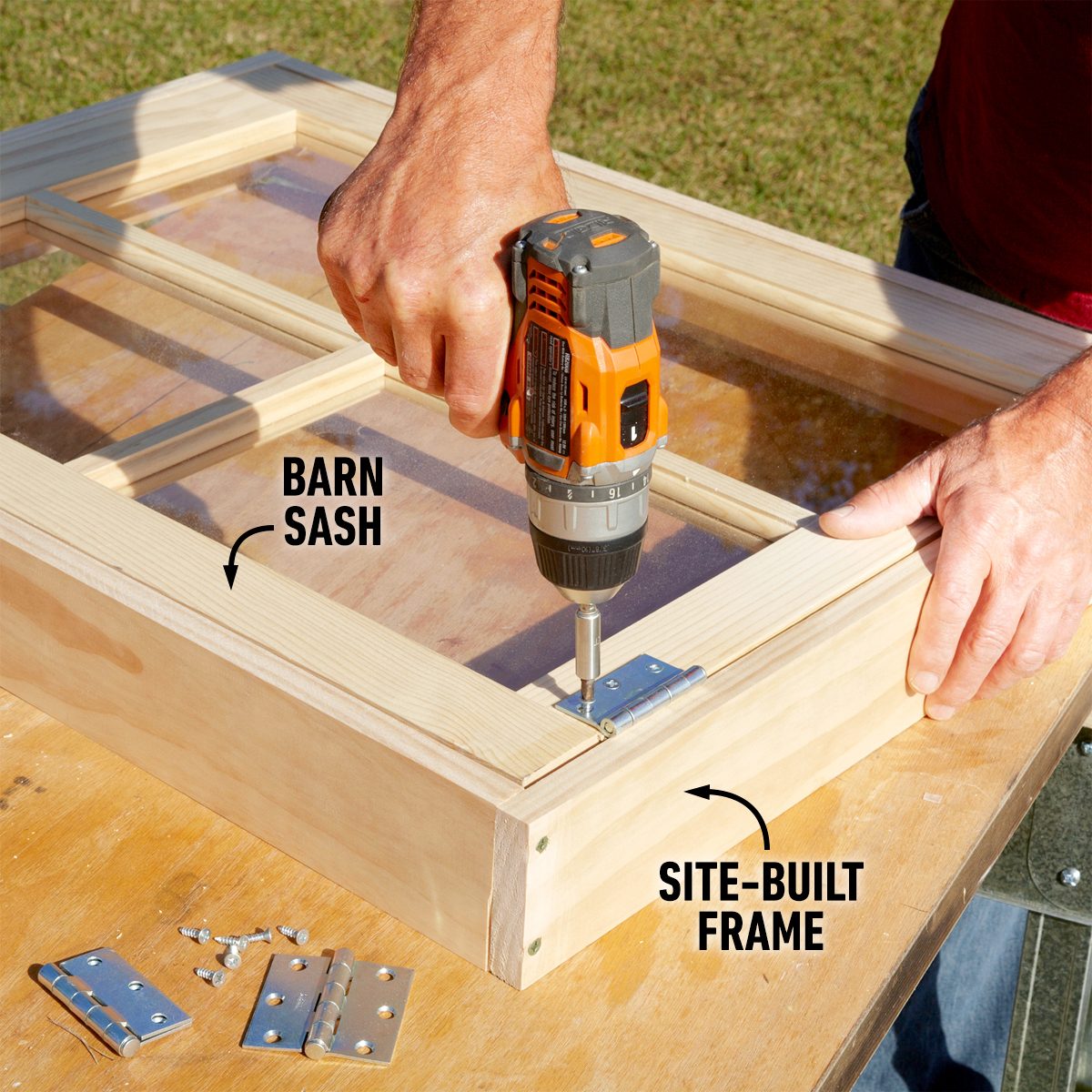 A hand uses a drill to attach hinges to a wooden frame, surrounded by screws and metal pieces, on a table in an outdoor setting.
