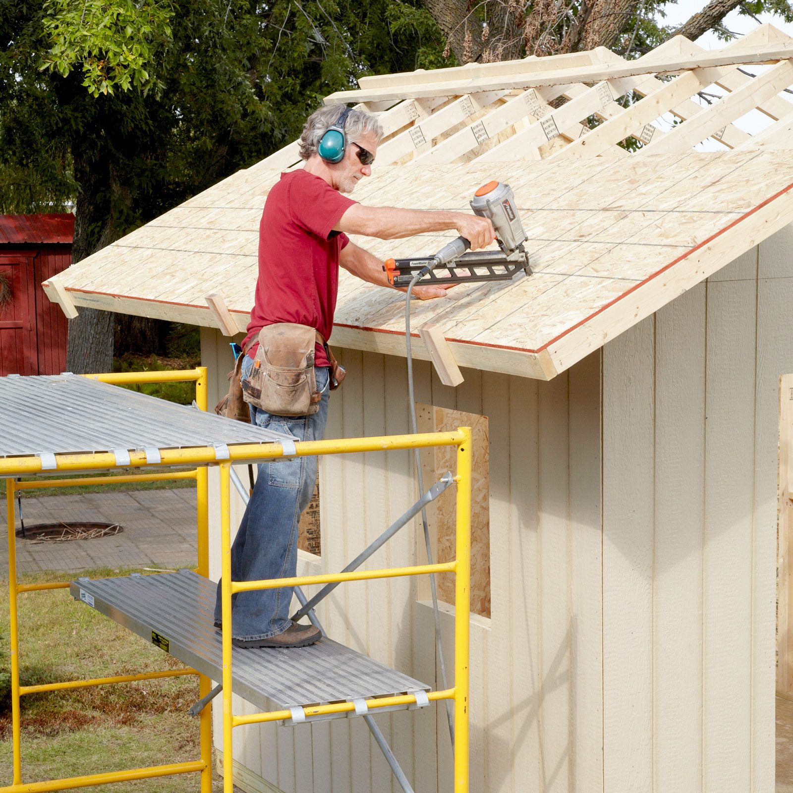 A man operates a nail gun on a roof, standing on scaffolding beside a partially constructed shed in a grassy outdoor environment.