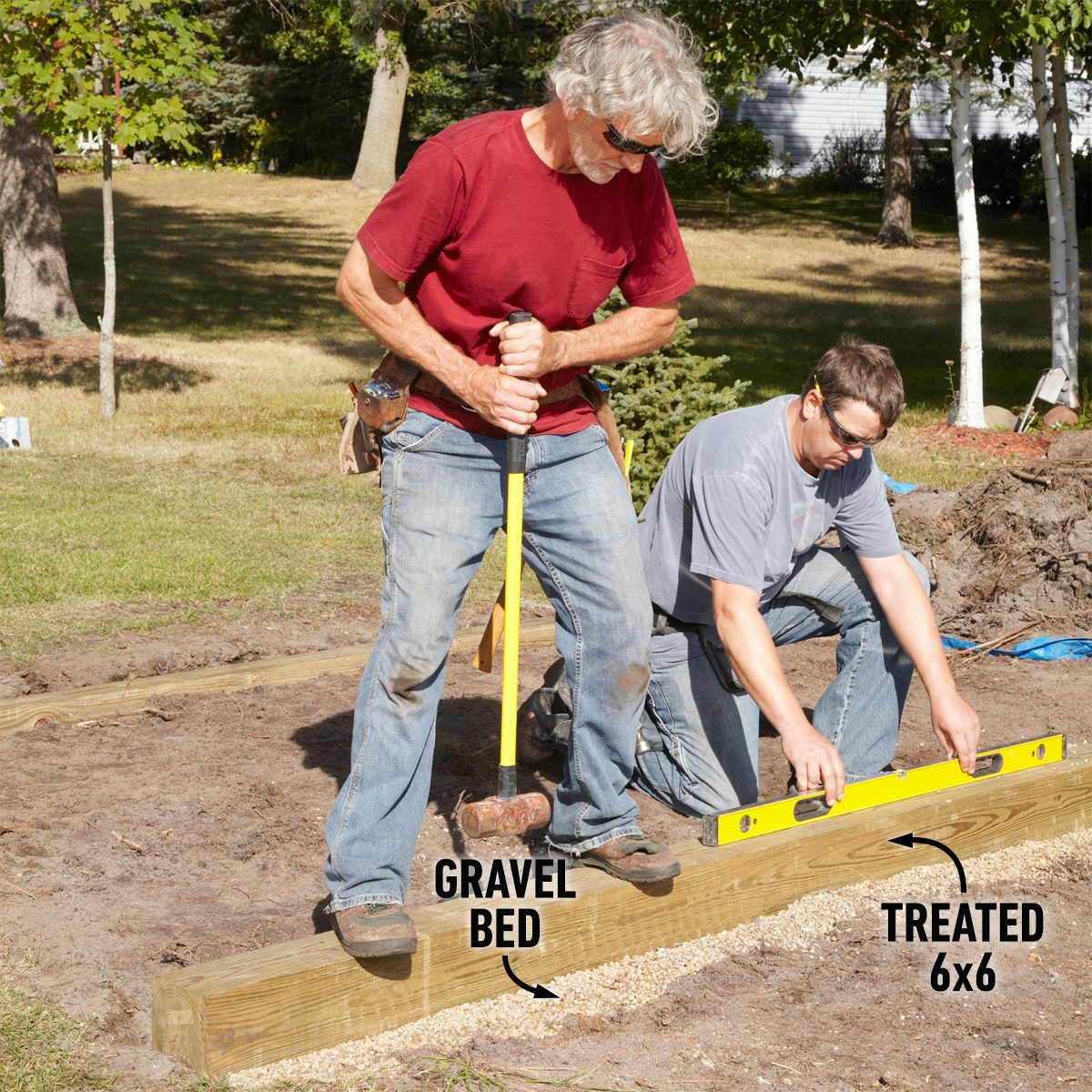 Two men work outdoors; one drives a stake into a gravel bed while the other levels a treated wood beam on the ground.
