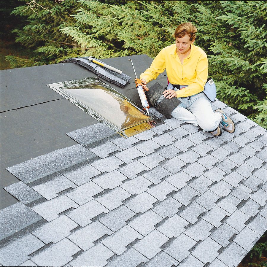 A woman is installing shingles on a roof while applying adhesive around a skylight, surrounded by trees. Tools are laid out nearby.