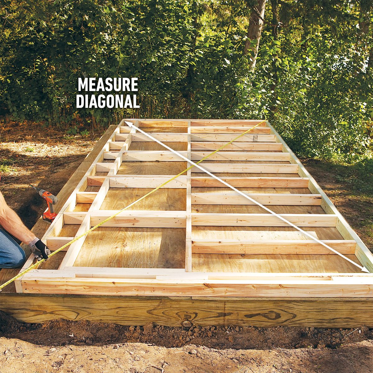 A person measures the diagonal of a wooden frame laid on the ground, surrounded by greenery, ensuring structural accuracy for construction.
