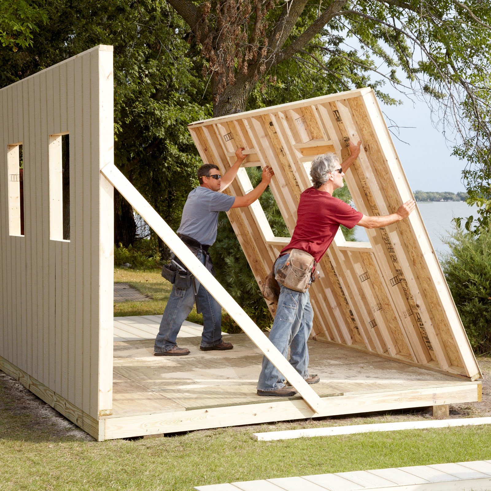 Two workers are raising a wall frame near a water body, surrounded by greenery, as they construct a building outdoors.