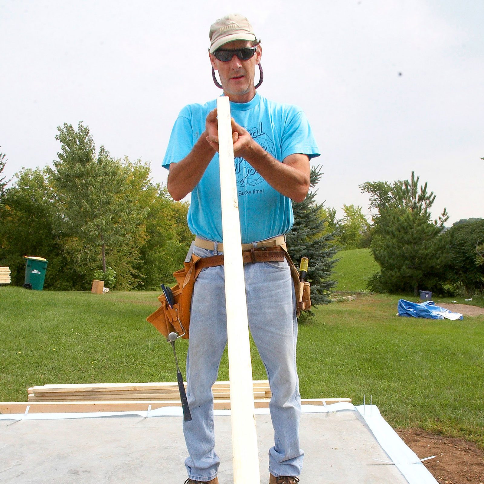 A man holds a long wooden board, focusing intently, on a grassy construction site surrounded by trees and construction materials.