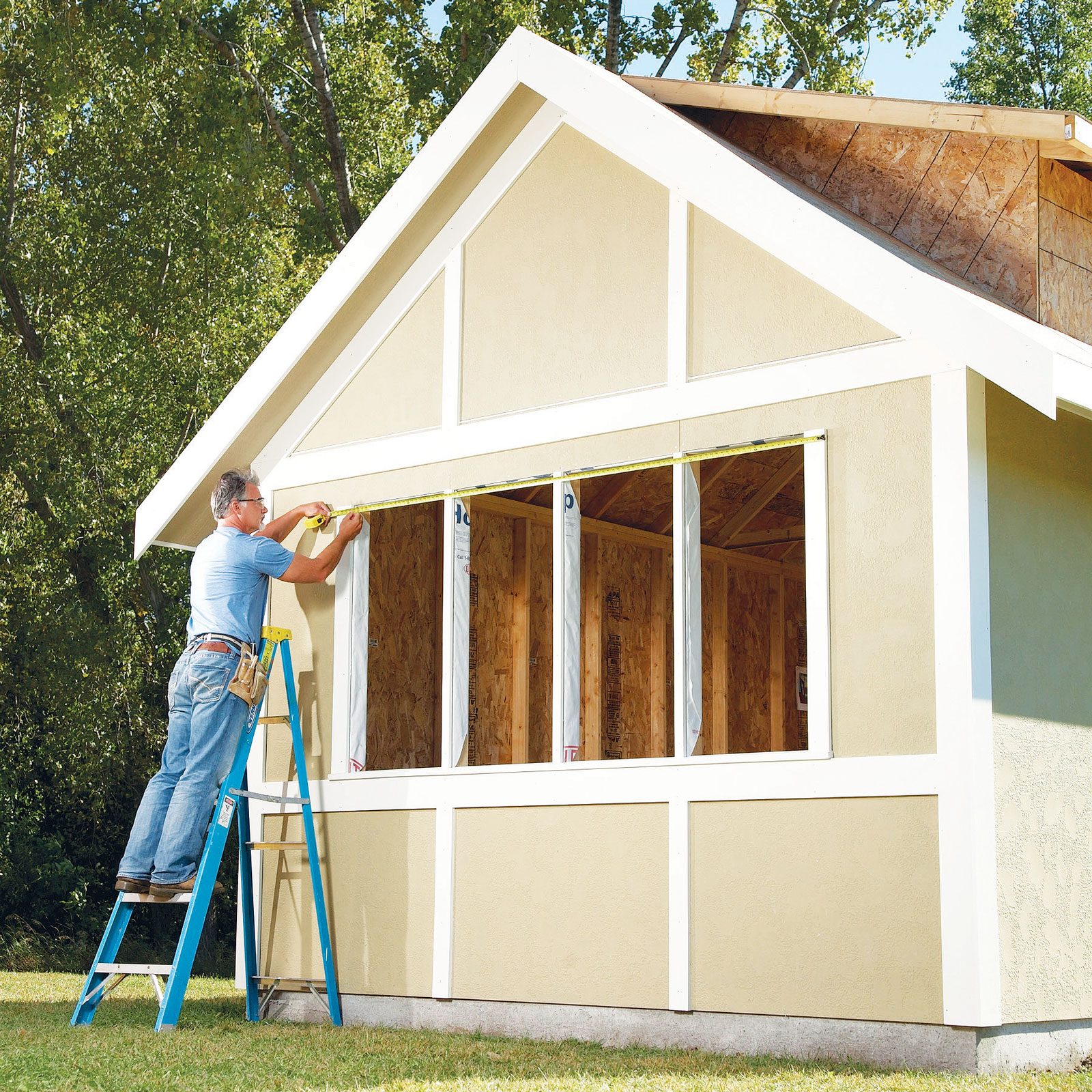 A man measures a window frame using a tape measure while standing on a ladder beside an unpainted house, surrounded by greenery.