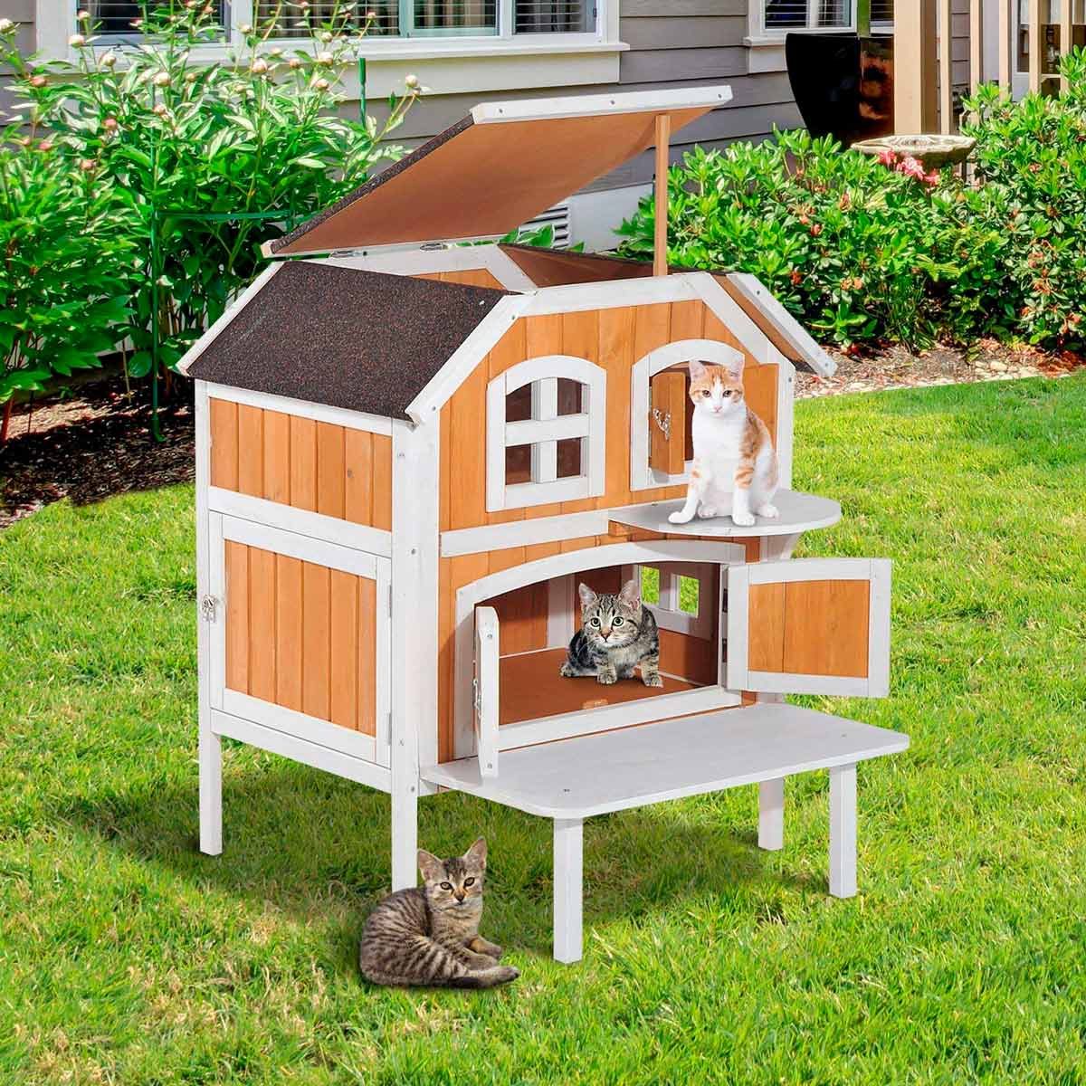 A wooden cat house sits on grass, with three cats exploring its levels, while a green shrub line and a house serve as the backdrop.