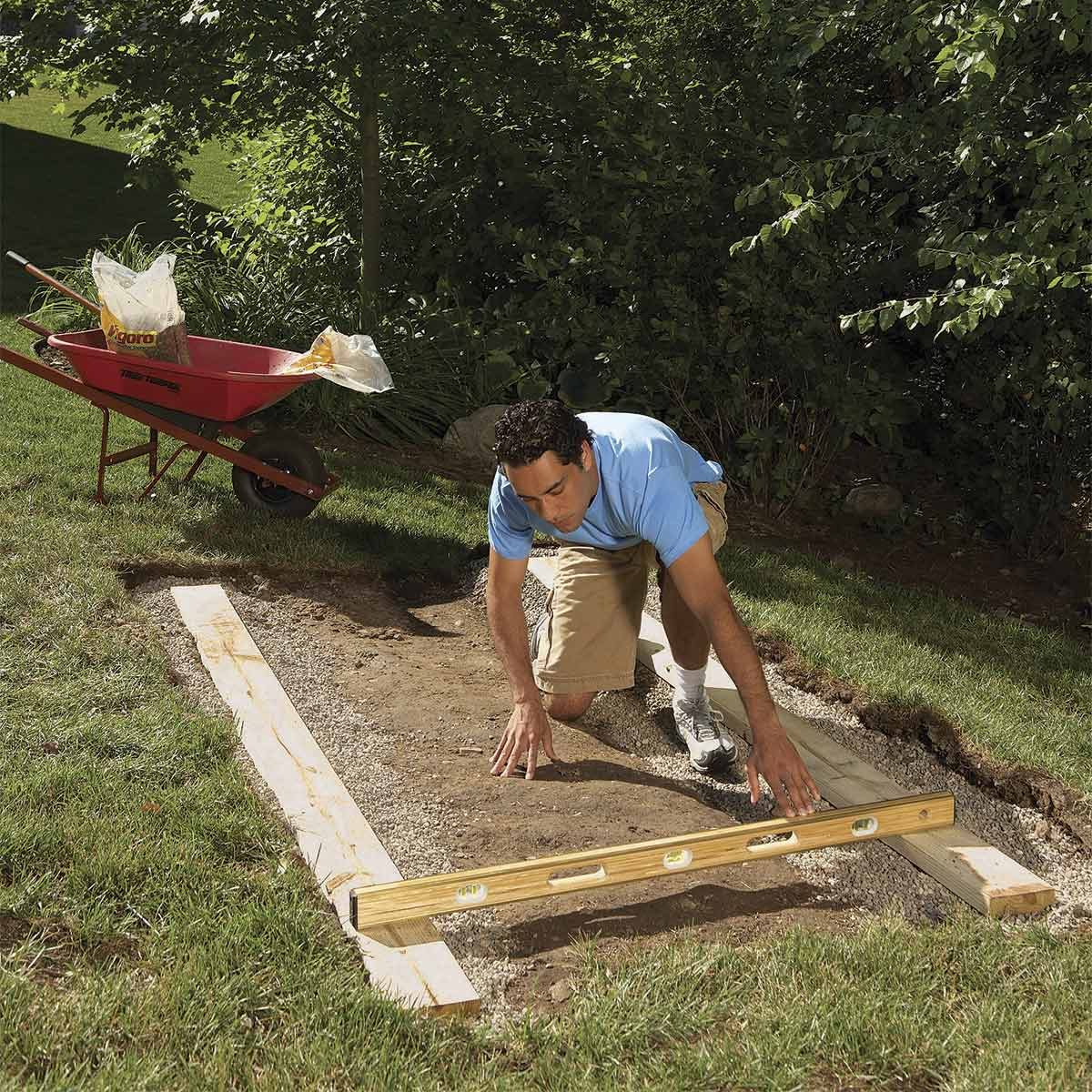 A man kneels on the ground, checking the level of a wooden board with a level tool, surrounded by a grassy yard and a wheelbarrow.
