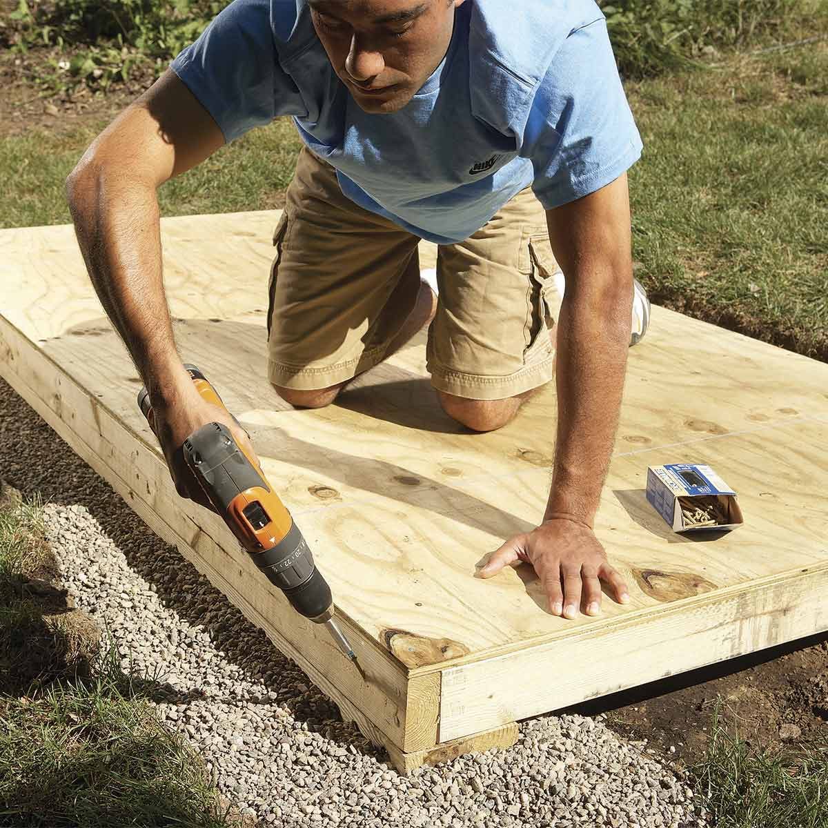 A man kneels on a wooden platform, using a drill to secure a board against gravel, surrounded by grass and tools.