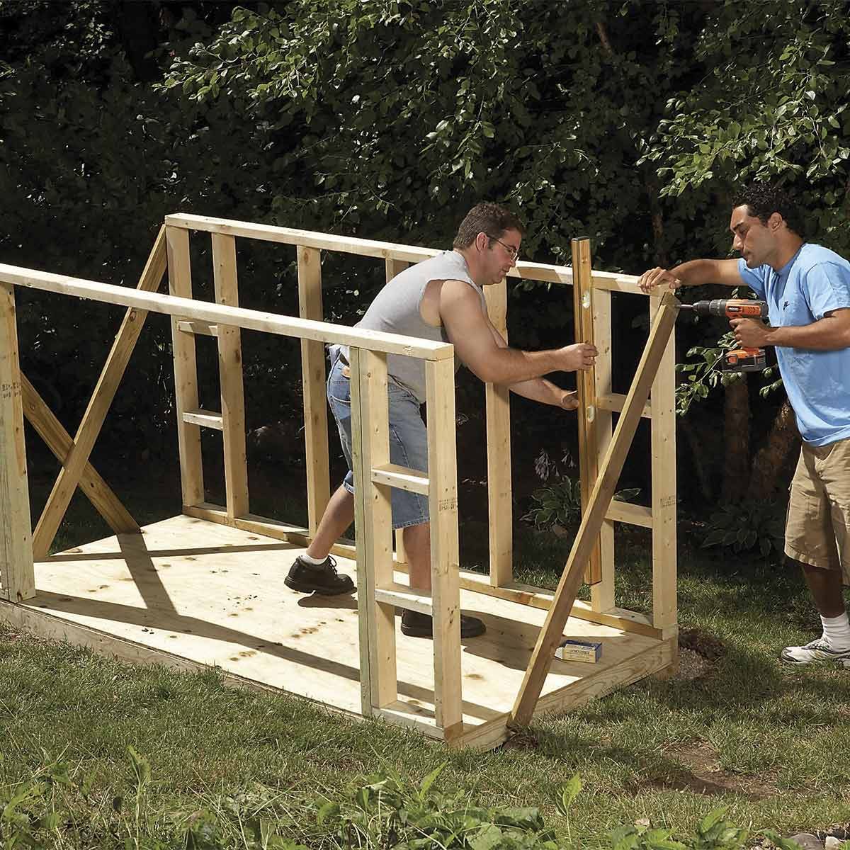 Two men construct a wooden frame for a structure in a grassy area, using tools and materials under a tree-filled backdrop.