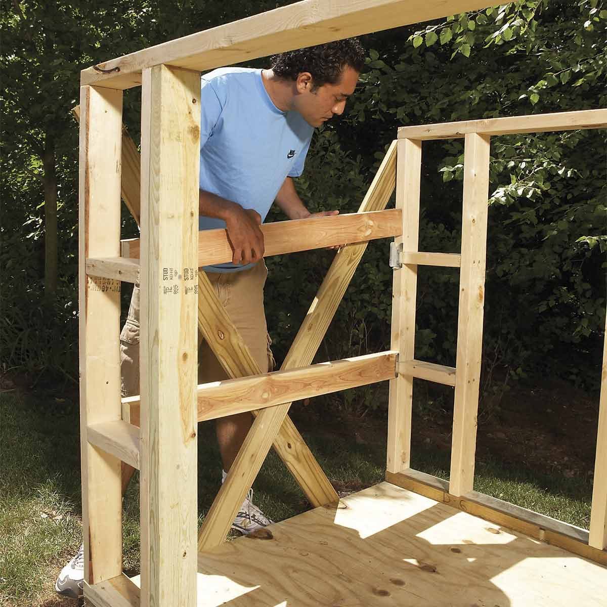 A person adjusts wooden frame beams while constructing a structure outdoors, surrounded by greenery and sunlight on a grassy surface.