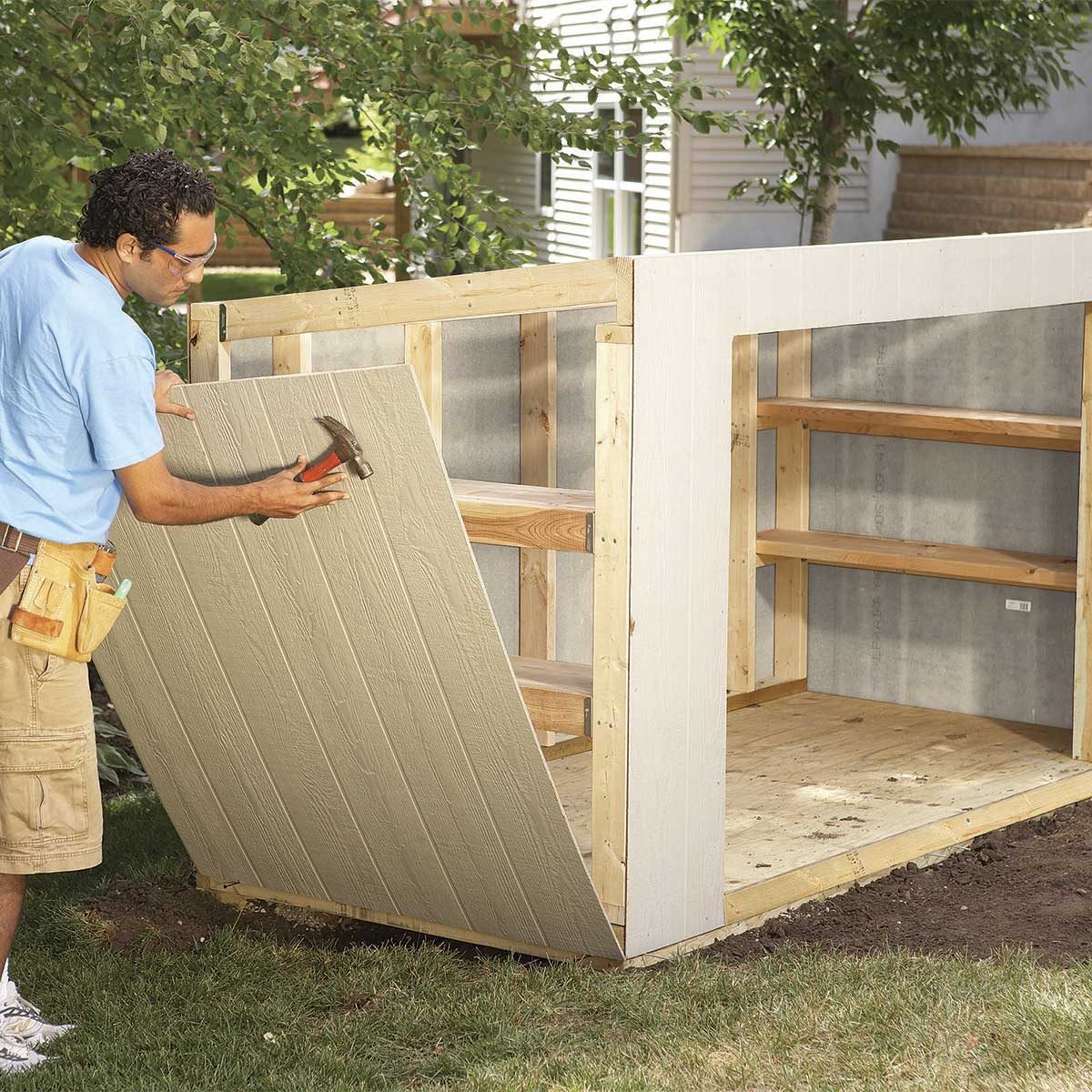 A person hammers a panel onto a partially constructed wooden shed in a grassy area, with shelves visible inside and trees in the background.