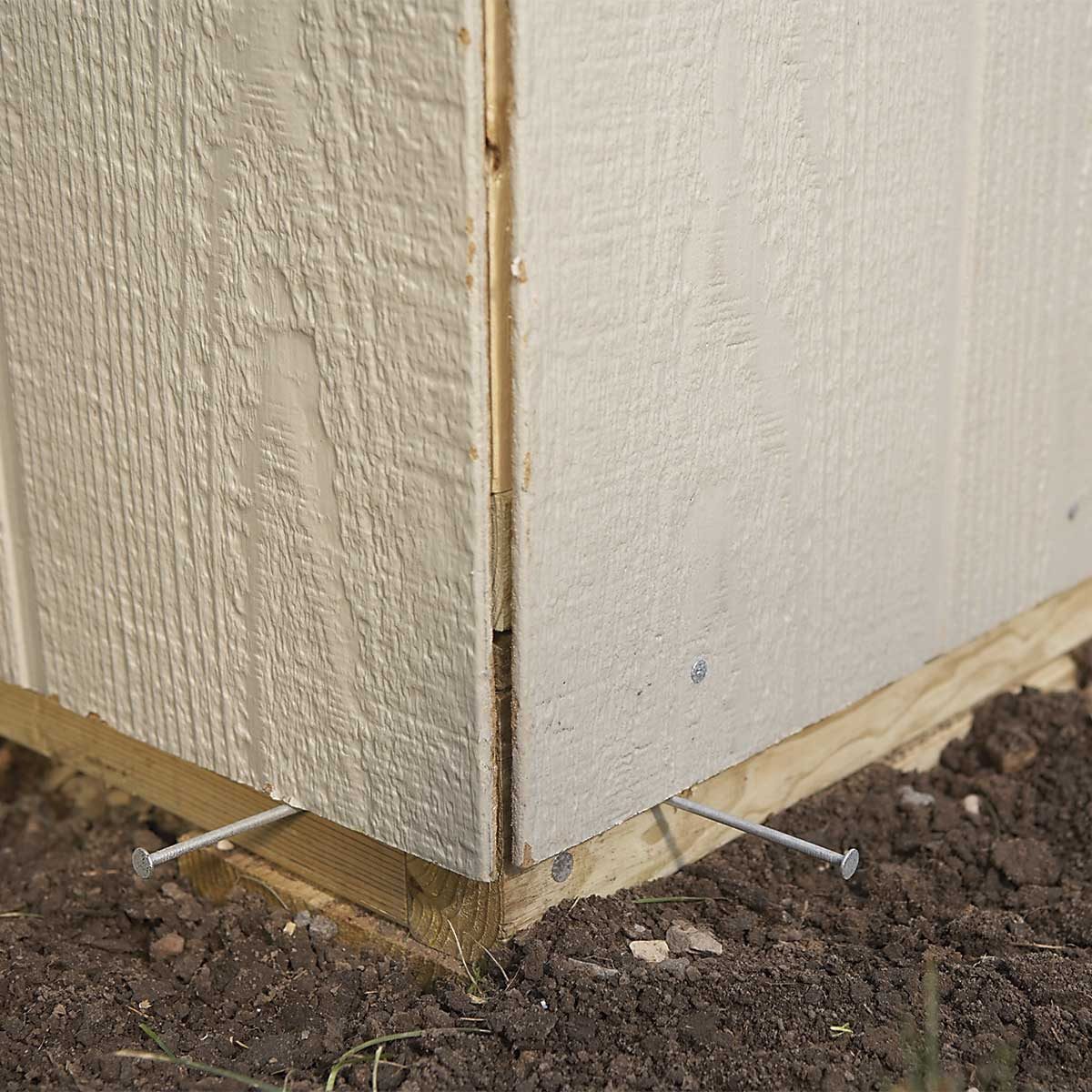 A wooden structure's corner is fastened to the ground with metal nails, surrounded by soil and grass. The panel shows wear and tear.