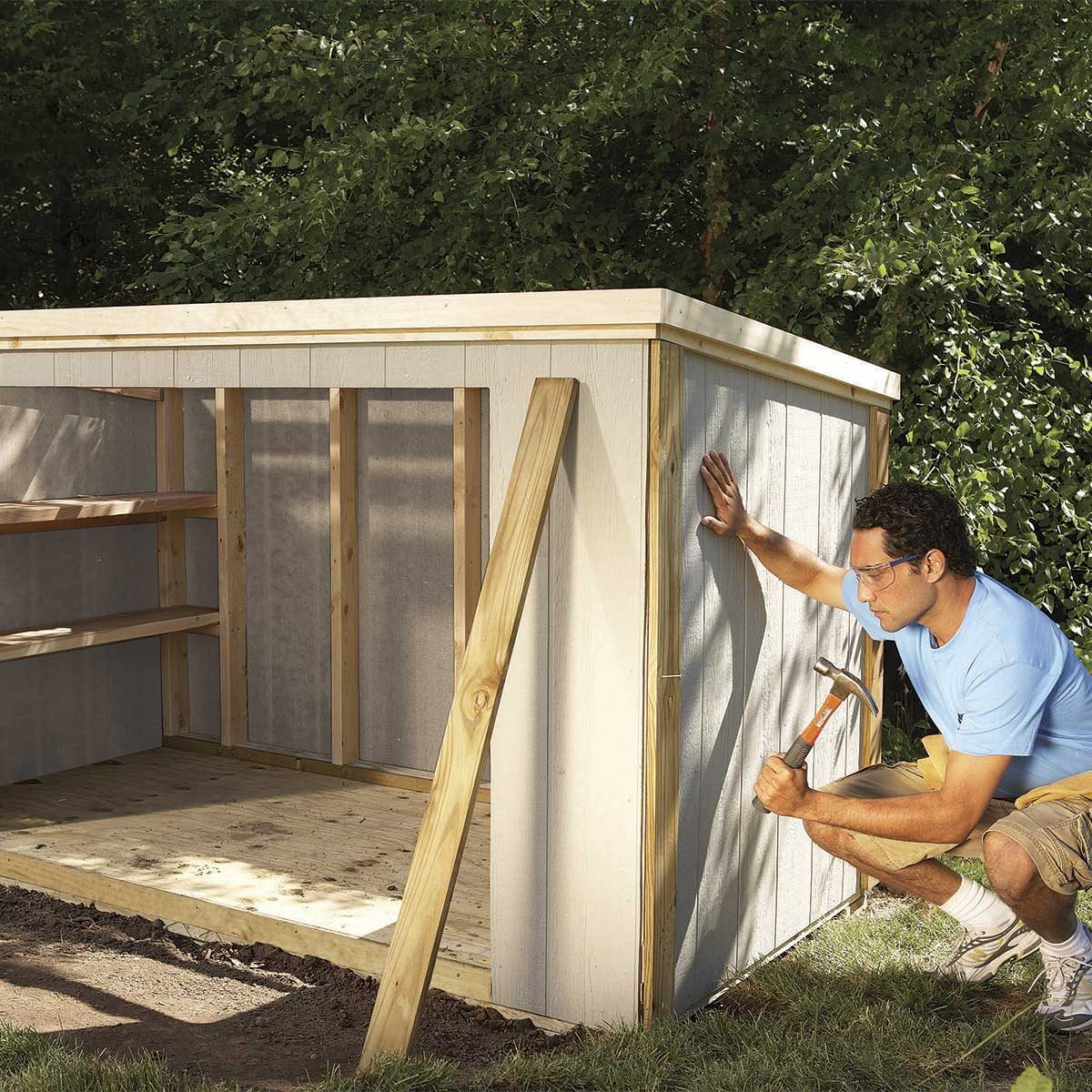 A man hammers a panel onto a wooden structure. The setting is an outdoor area surrounded by trees, with shelves visible inside the building.