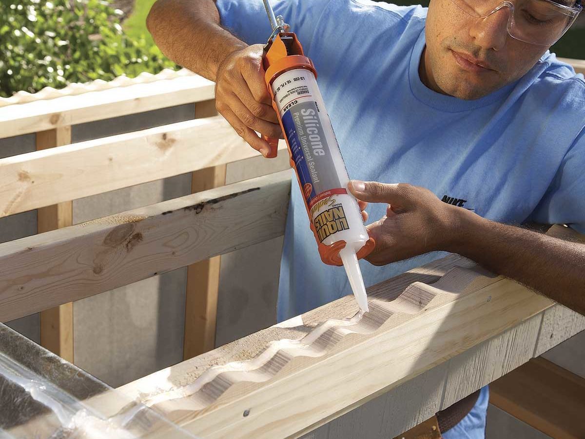 A man applies silicone sealant using a caulking gun on a wooden surface, with green grass and a structure visible in the background.