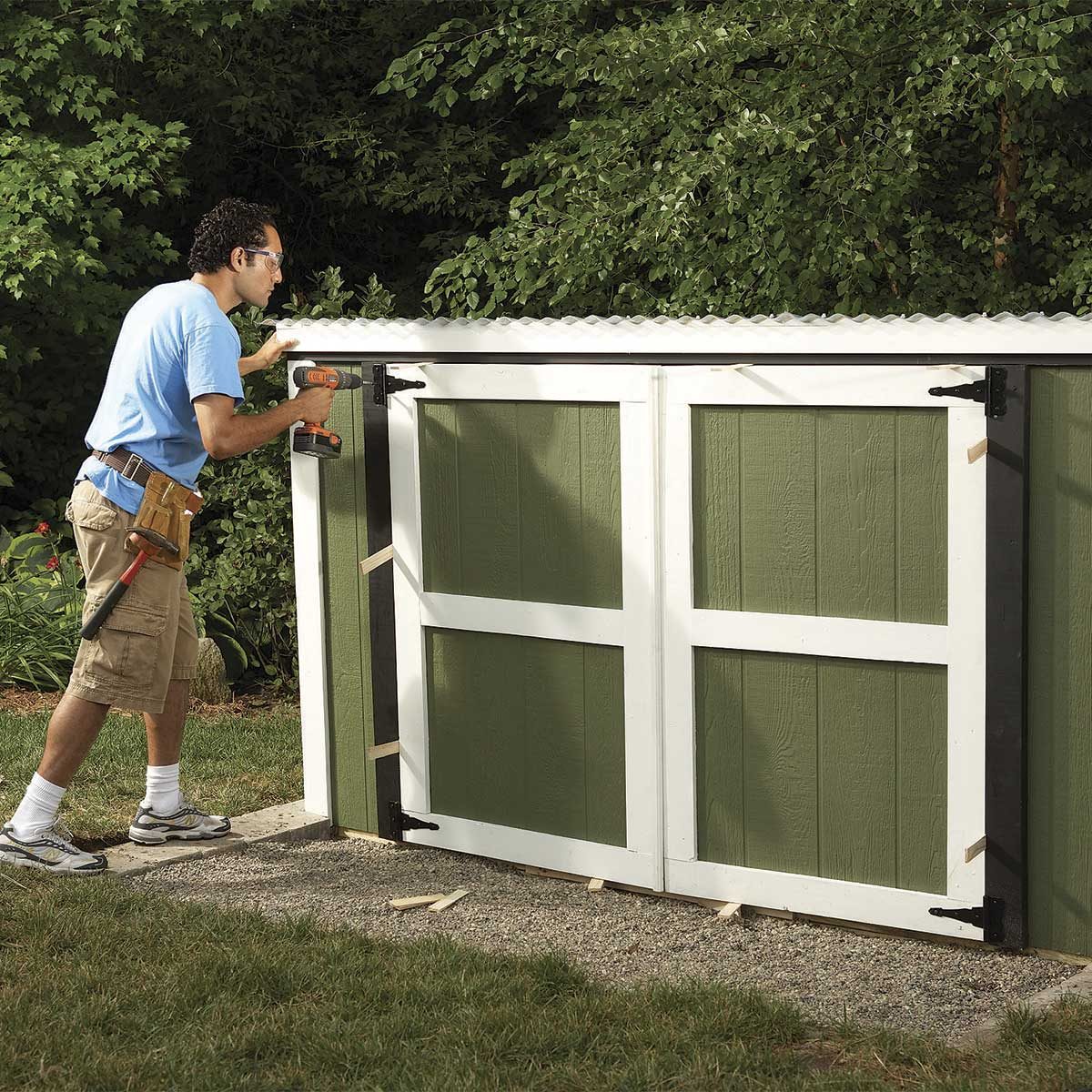 A man uses a power tool to attach a door frame to a green shed, surrounded by greenery and gravel, during a home improvement project.