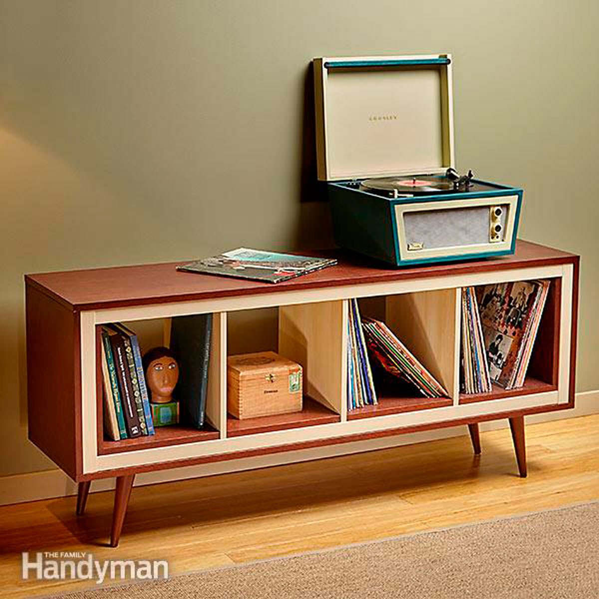 A vintage record player sits on a wooden console, surrounded by vinyl records, books, and a small decorative box against a muted wall.