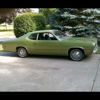 A green vintage car is parked on a concrete driveway, surrounded by lush bushes and trees under a clear sky.