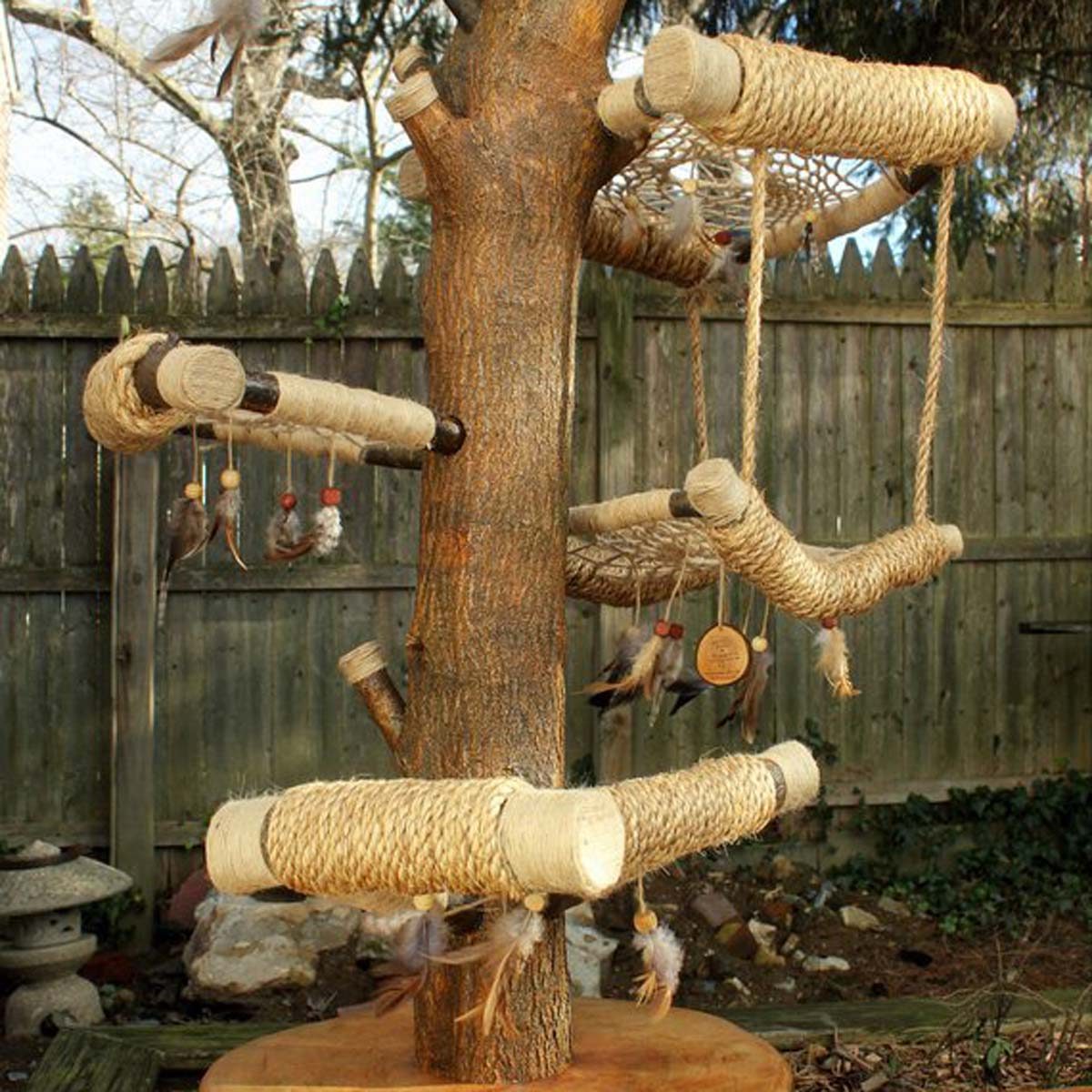 A wooden tree structure features multiple rope-wrapped perches and hanging toys, situated in a garden with a wooden fence and natural ground cover.