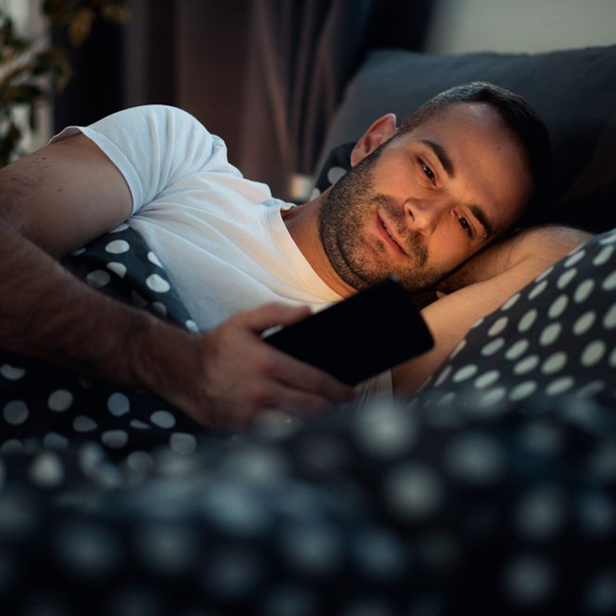 A man lies in bed, holding a smartphone and looking at it intently, surrounded by patterned bedding in a softly lit room.