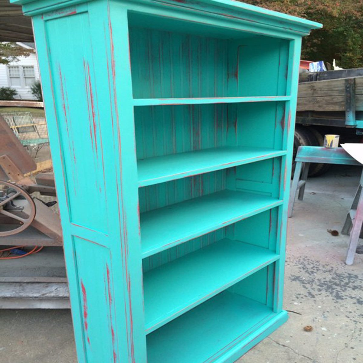 A turquoise wooden bookshelf stands upright, featuring five open shelves, outdoors with a blurred background of tools and an unfinished wooden structure.