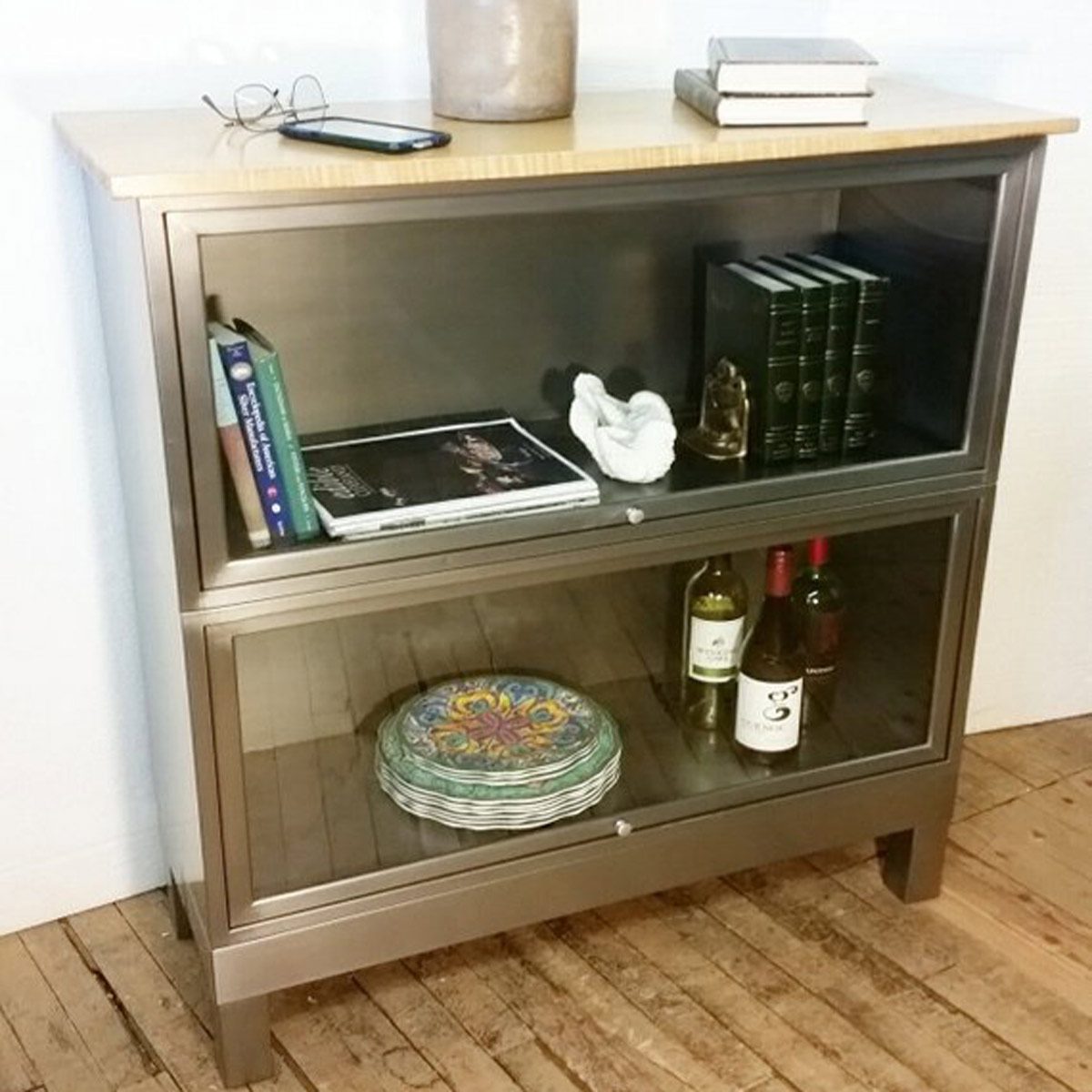 A modern cabinet displays books, a sculpture, and bottles of wine, resting on a wooden floor, while glasses and a phone are on top.