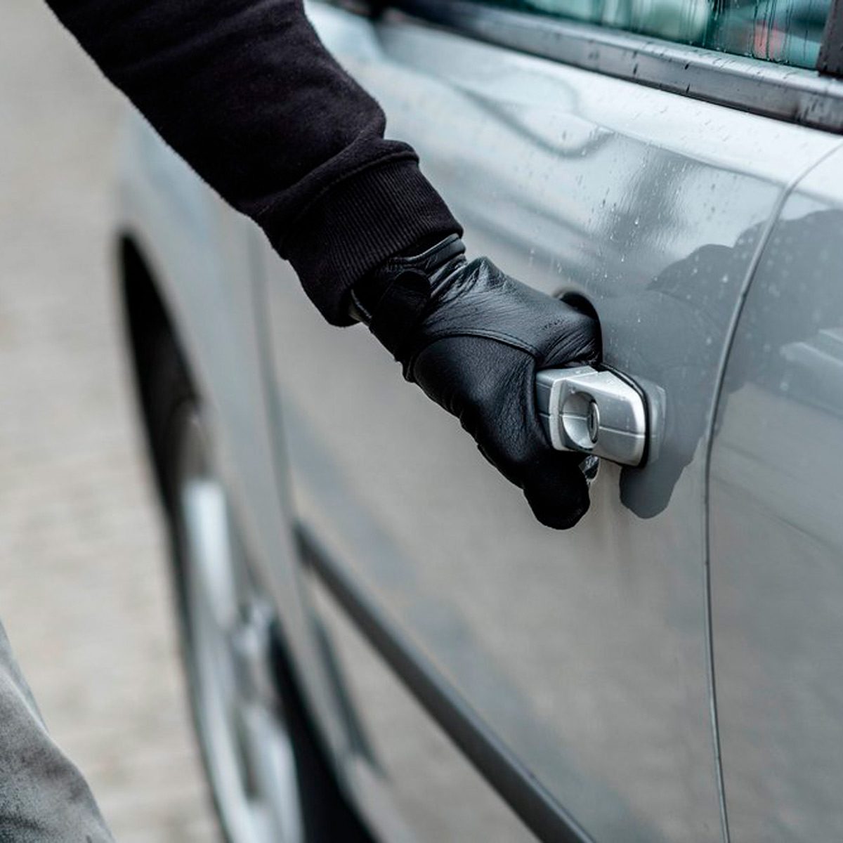 A gloved hand is pulling the door handle of a gray car, suggesting an attempt to gain entry in an outdoor setting.