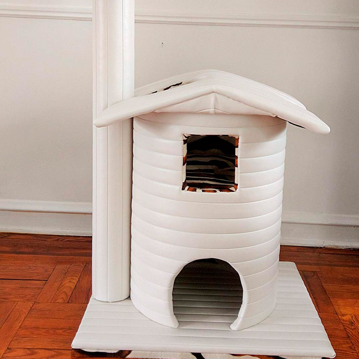 A white, cylindrical pet house sits on a wooden floor with a tall scratching post nearby, providing a cozy space for a pet to explore.