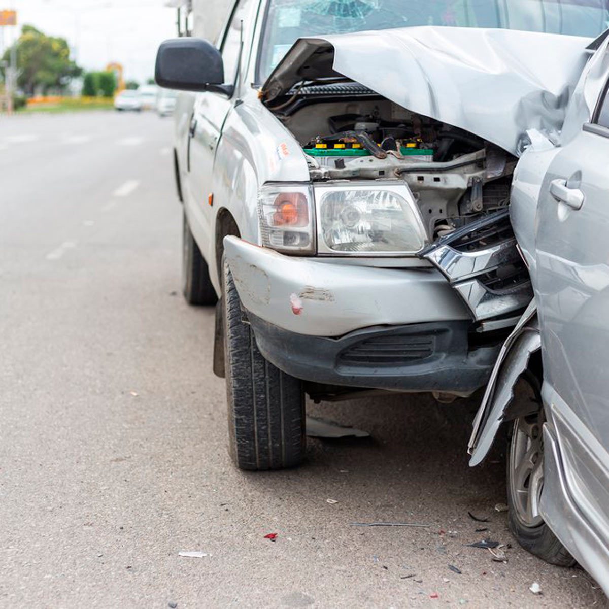 A damaged silver pickup truck is parked with its hood open, showing engine components, while a silver car has a crumpled front, situated on a road.