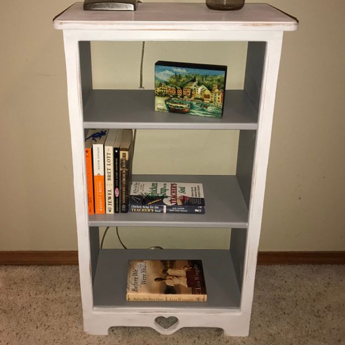 A white bookshelf holds several books and a small painting, situated against a plain wall with carpet below. A lamp rests on top.